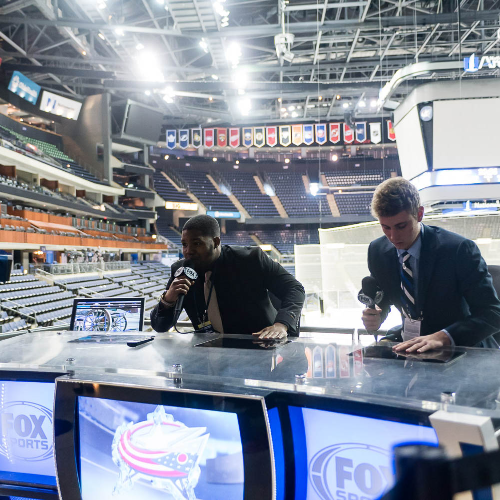 Stepping behind the curtain during their job shadow of the Columbus Blue Jackets, two students sit at a news table