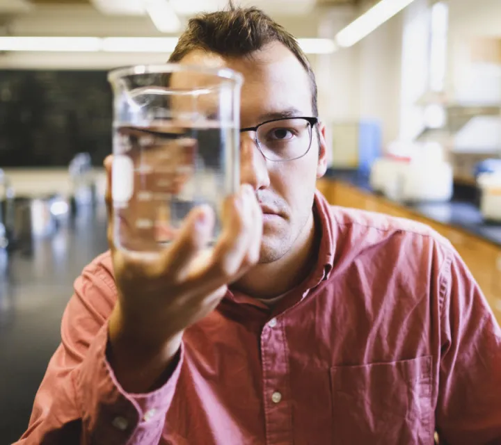 Marietta College student Joe Tucker '21 holds a graduated cylinder in front of half of his face for a portrait