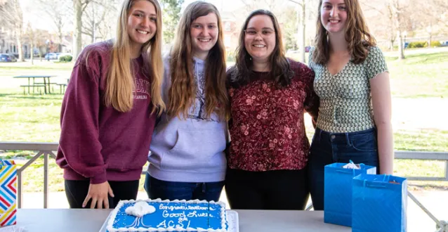 Four women students standing behind a cake