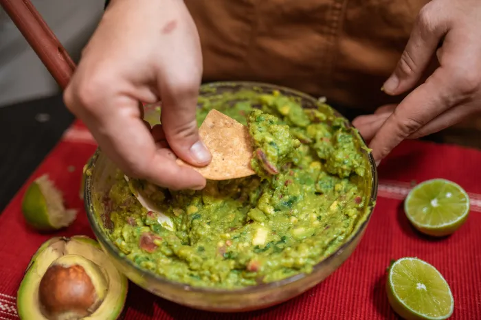 A hand, holding a tortilla chip, dipping into a bowl of guacamole.