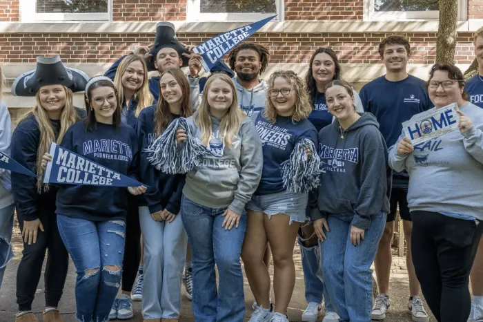16 Marietta College students stand together, all smiling, wearing navy blue, gray, and white Marietta College attire, holding Marietta College flags and signs