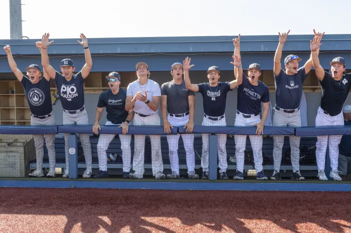 Marietta College baseball players, wearing navy and white uniforms stand together in the dugout cheering. 