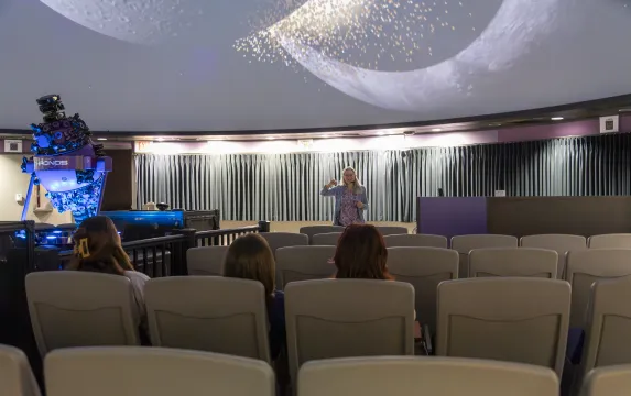 Professor Ann Bragg presents in the newly renovated planetarium. Three students sit in the audience. 