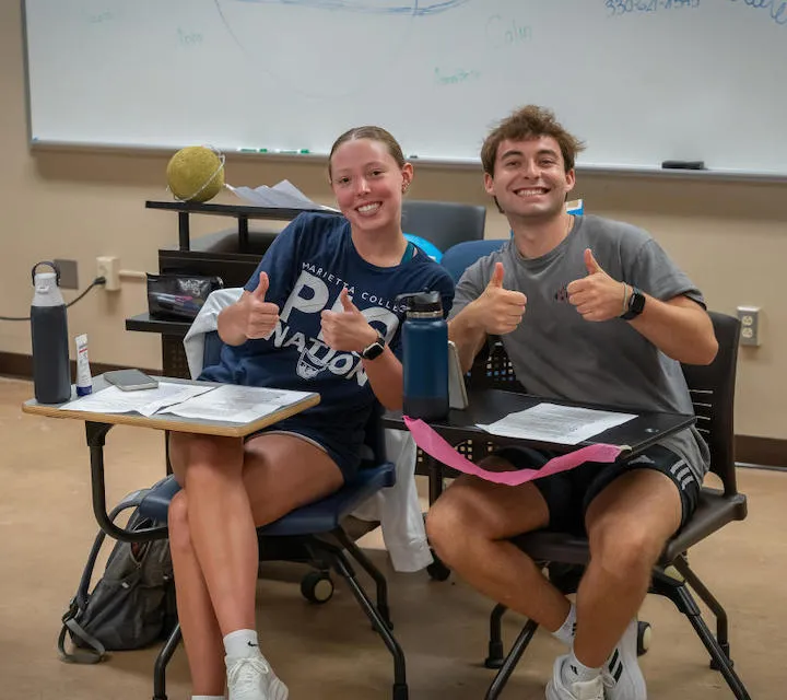 Two Marietta College students smile and give thumbs up while sitting in a classroom