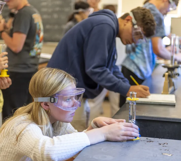 Female chemistry student measuring liquid in a beaker; students in the background taking lab notes