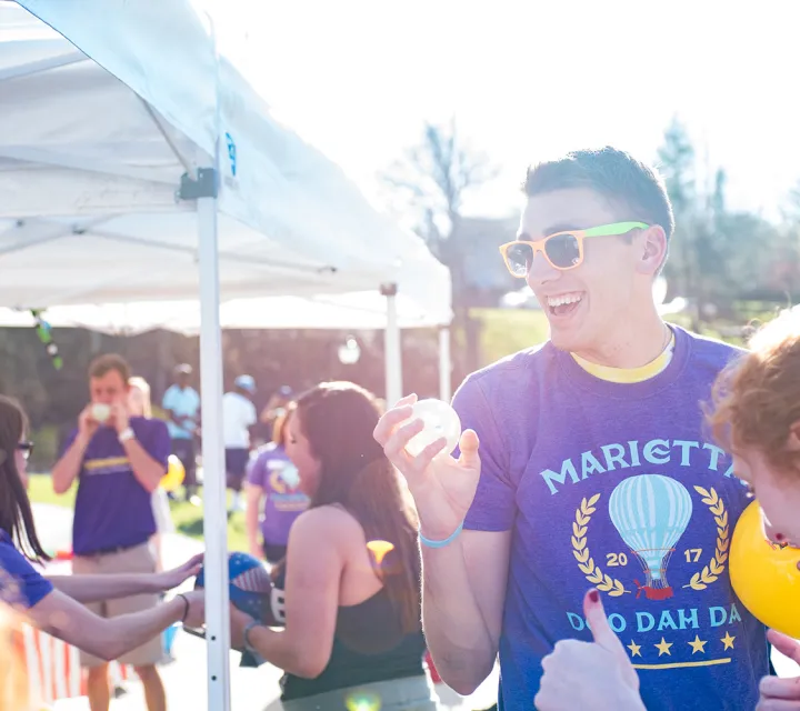 Male student wearing purple Doo Dah Day shirt gets ready to throw whiffle ball at carnival game