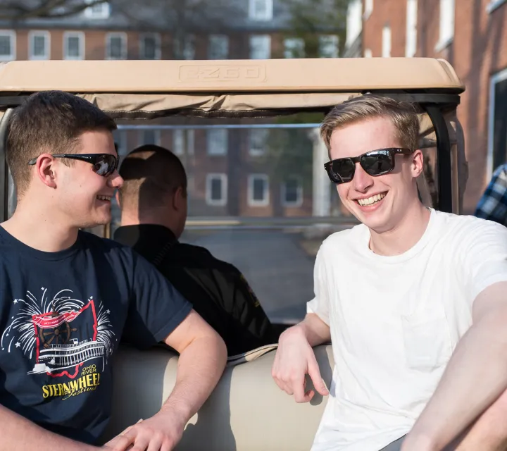 Two students sit on the back of a golf cart and smile