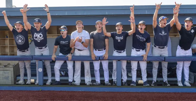 Marietta College baseball players, wearing navy and white uniforms stand together in the dugout cheering. 