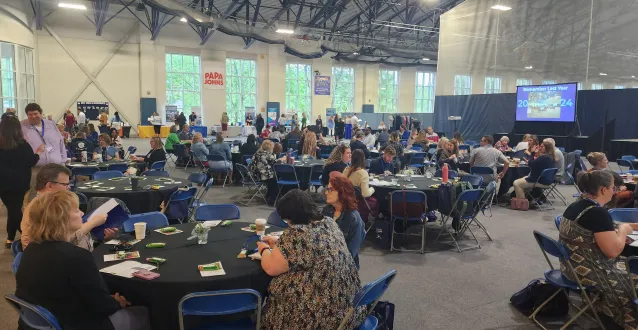 Attendees sit at round tables in the Dyson Baudo Recreation Center during the 2024 NonProfits LEAD Conference as a speaker stands on stage at the podium