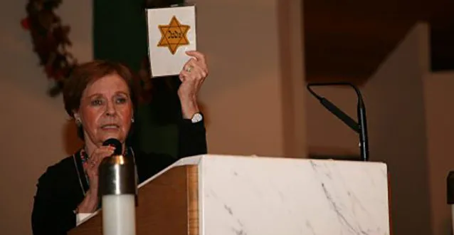 Marion Blumenthal Lazan speaking behind a podium and holding up a Jewish star