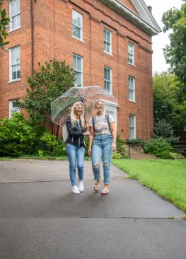 Two students walking in the rain while holding an umbrella in front of Erwin Hall on the Marietta College Campus